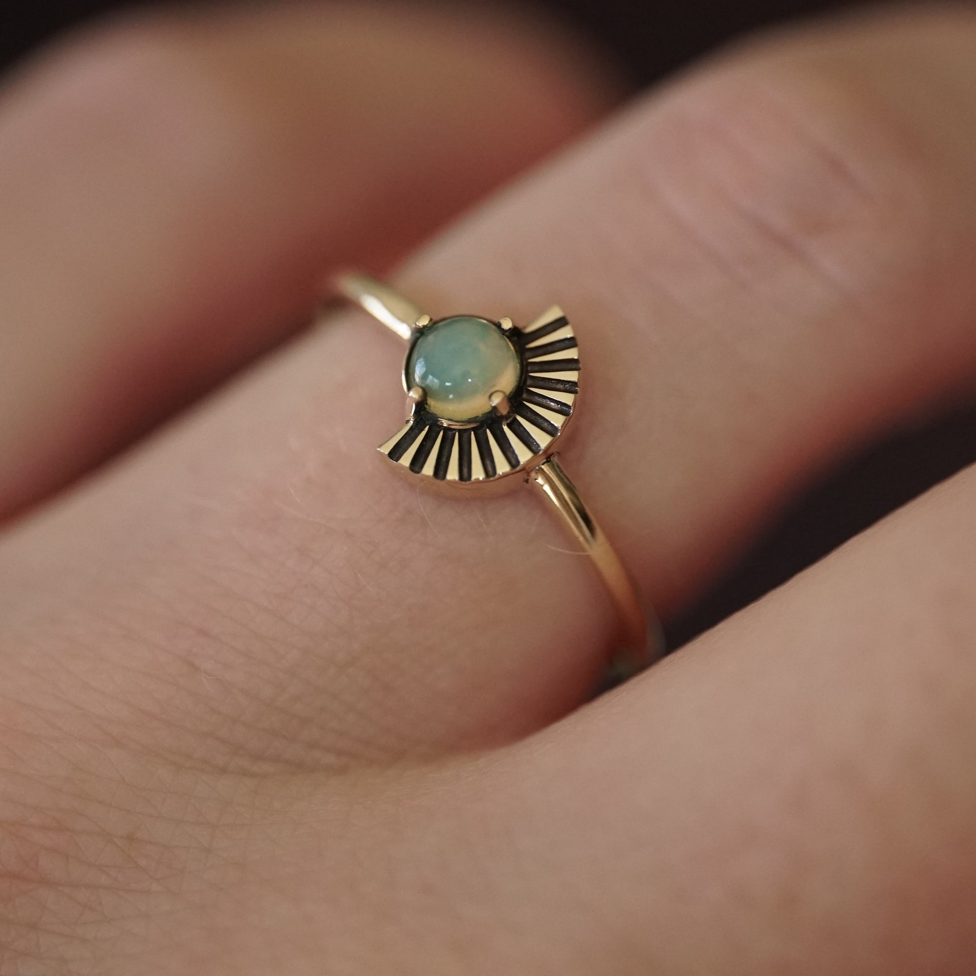 a cabochon cut opal in a yellow gold ring with an art deco fan element to one side is shown on a light-skinned left hand