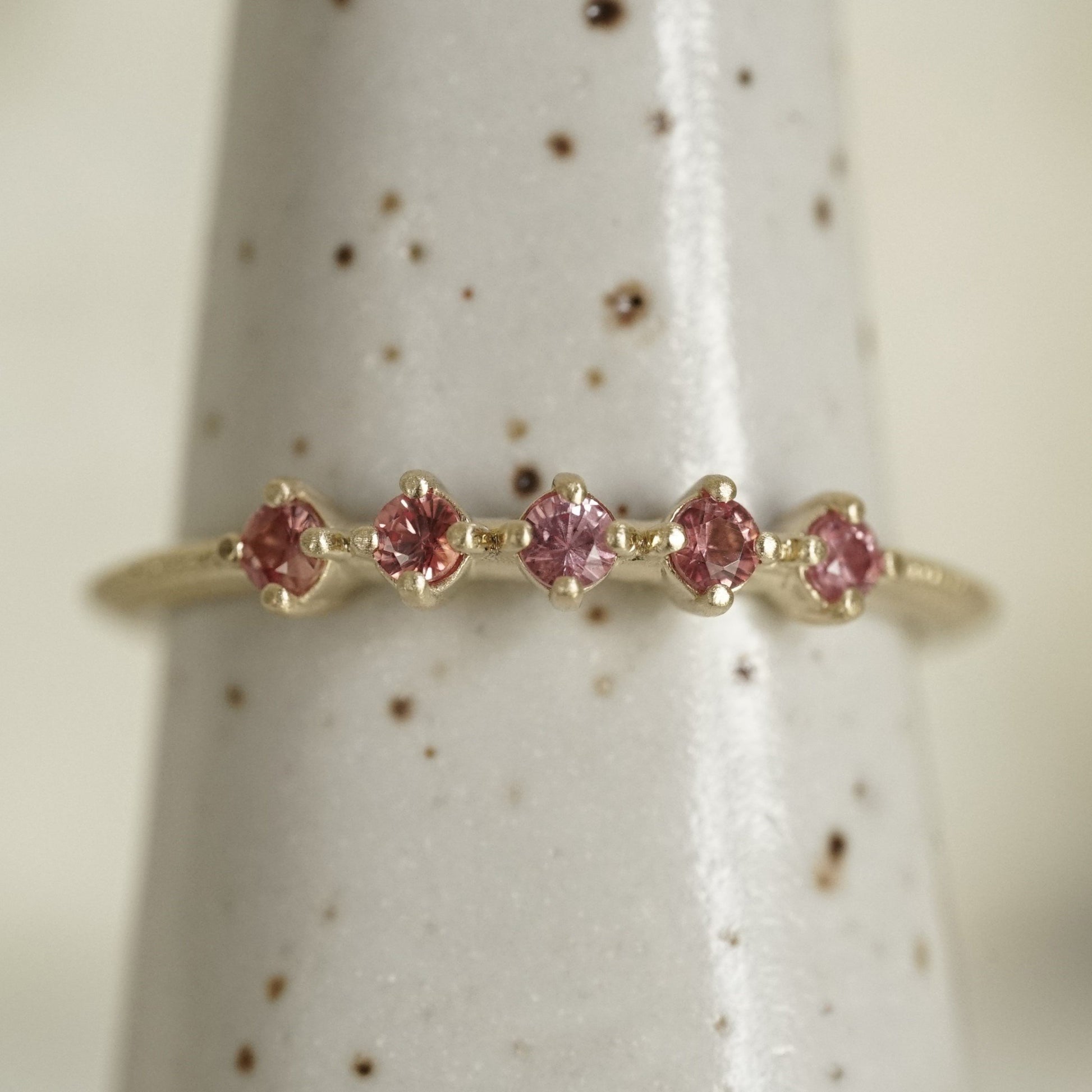a yellow gold band with 5 prong set pink padparadscha sapphires in prong settings on a white ceramic display