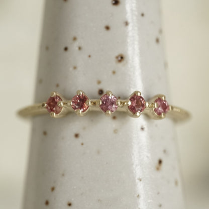 a yellow gold band with 5 prong set pink padparadscha sapphires in prong settings on a white ceramic display