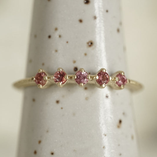 a yellow gold band with 5 prong set pink padparadscha sapphires in prong settings on a white ceramic display