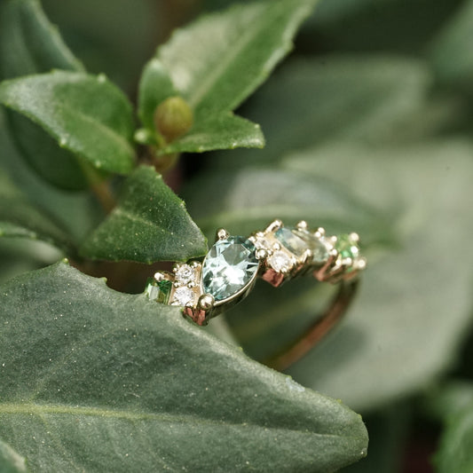 a yellow gold ring features green and teal gemstones paired with round white diamonds in a playful design resting in green foliage