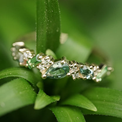 a macro photo of a yellow gold ring features green and teal gemstones paired with round white diamonds in a playful design on a green plant