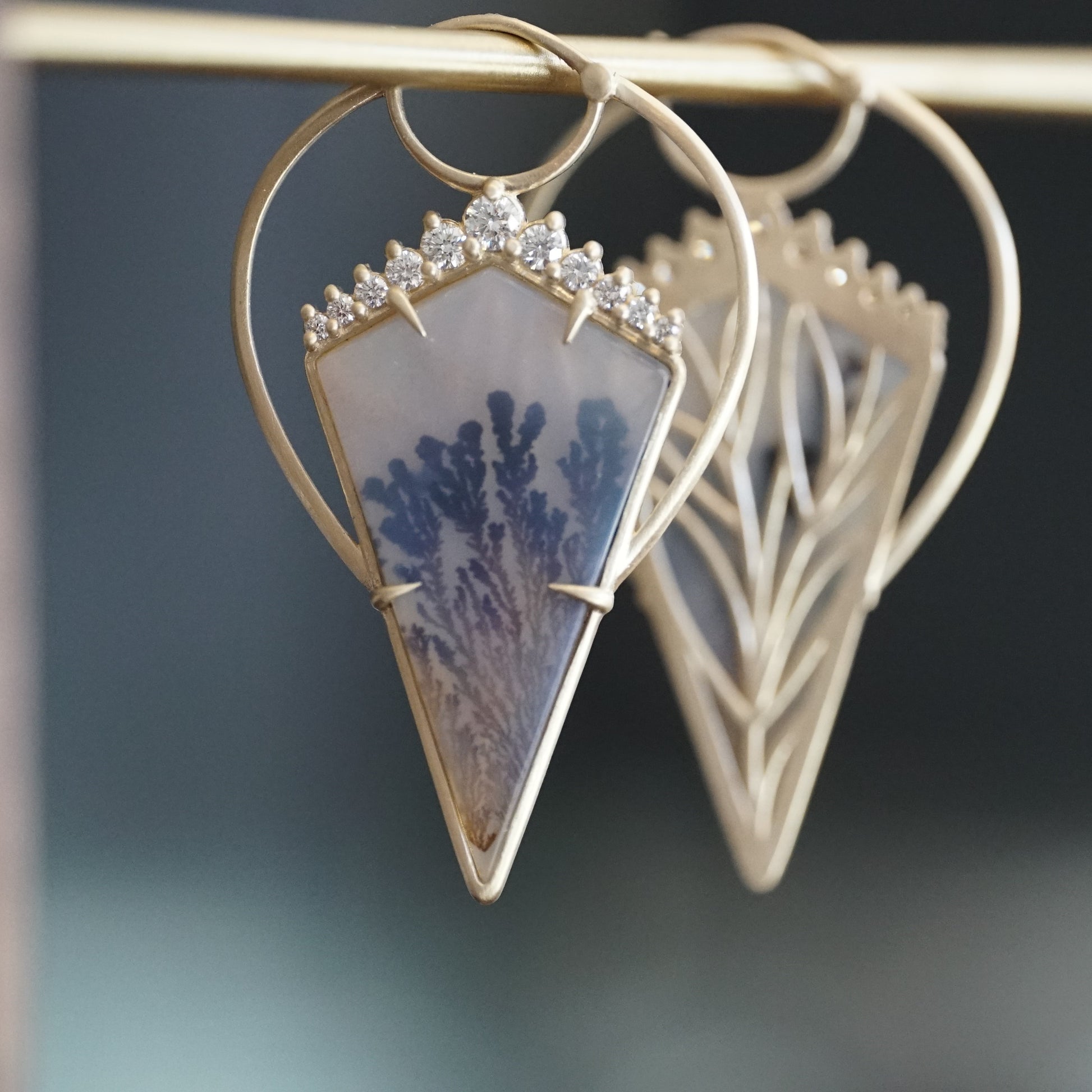 a pair of yellow gold earrings with kite shape dendritic agates and round diamonds hangs on a brass bar