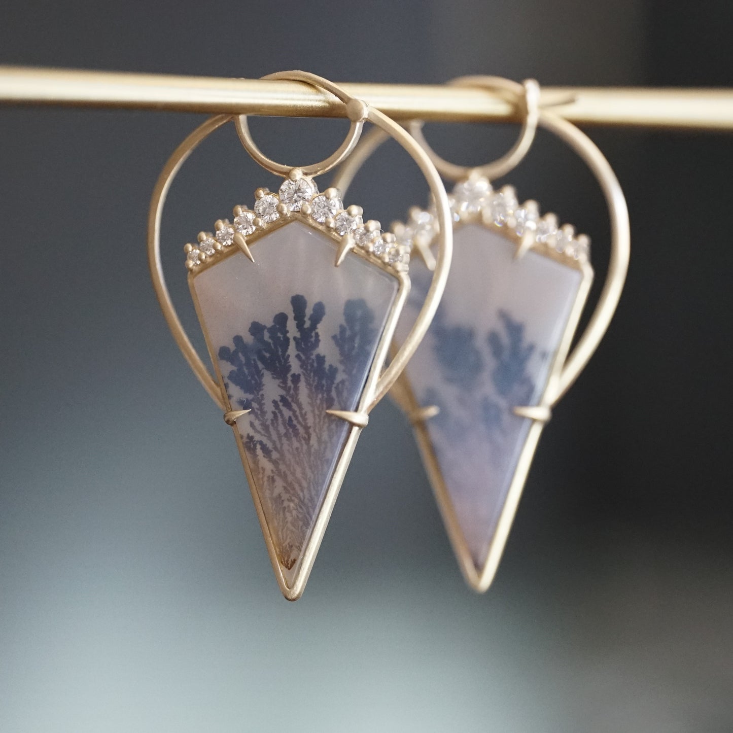 a pair of yellow gold earrings with kite shape dendritic agates and round diamonds hangs on a brass bar