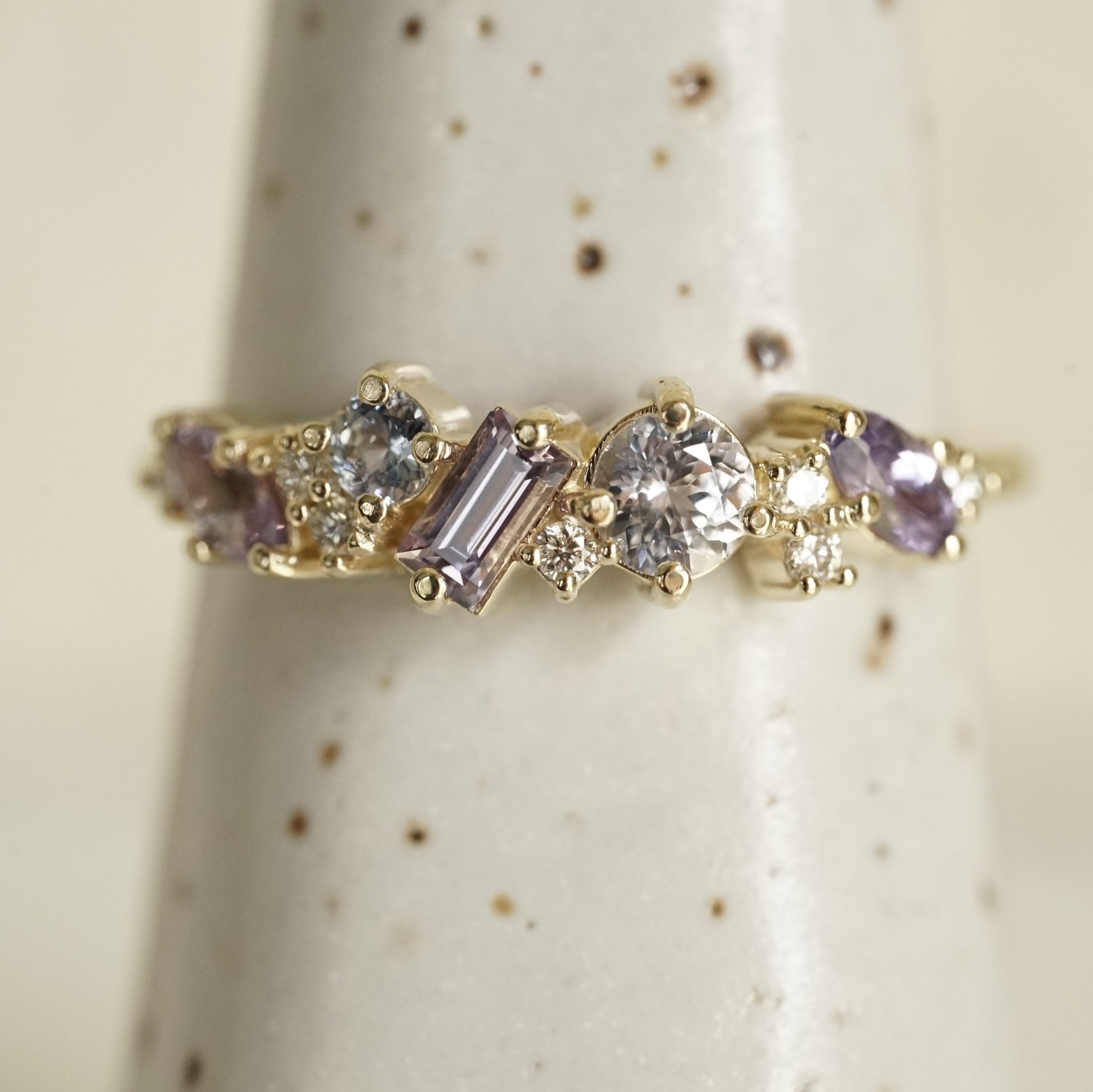 a yellow gold ring featuring purple and blue sapphires of mixed shapes and sizes are paired with round diamonds in a playful design resting on a white ceramic display