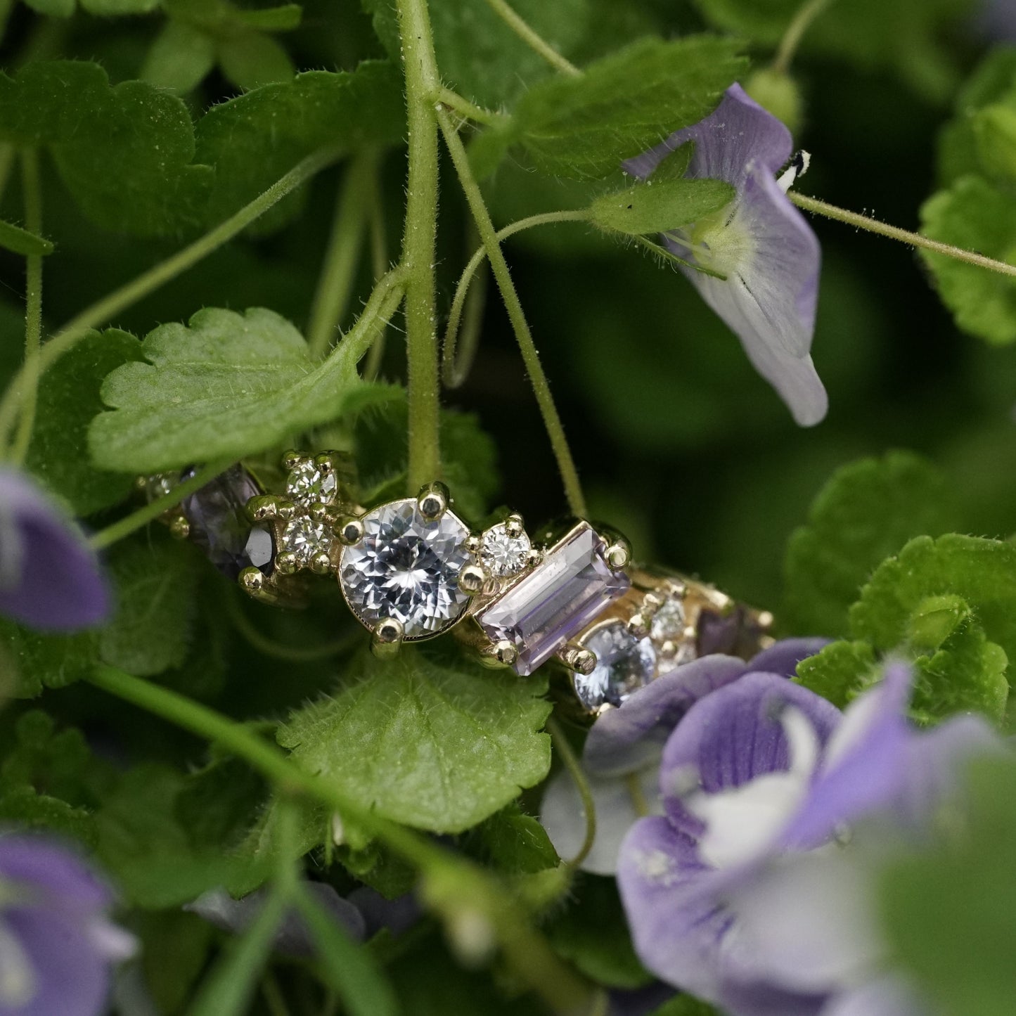 a yellow gold ring featuring purple and blue sapphires of mixed shapes and sizes are paired with round diamonds in a playful design. the ring rests in green and purple foliage