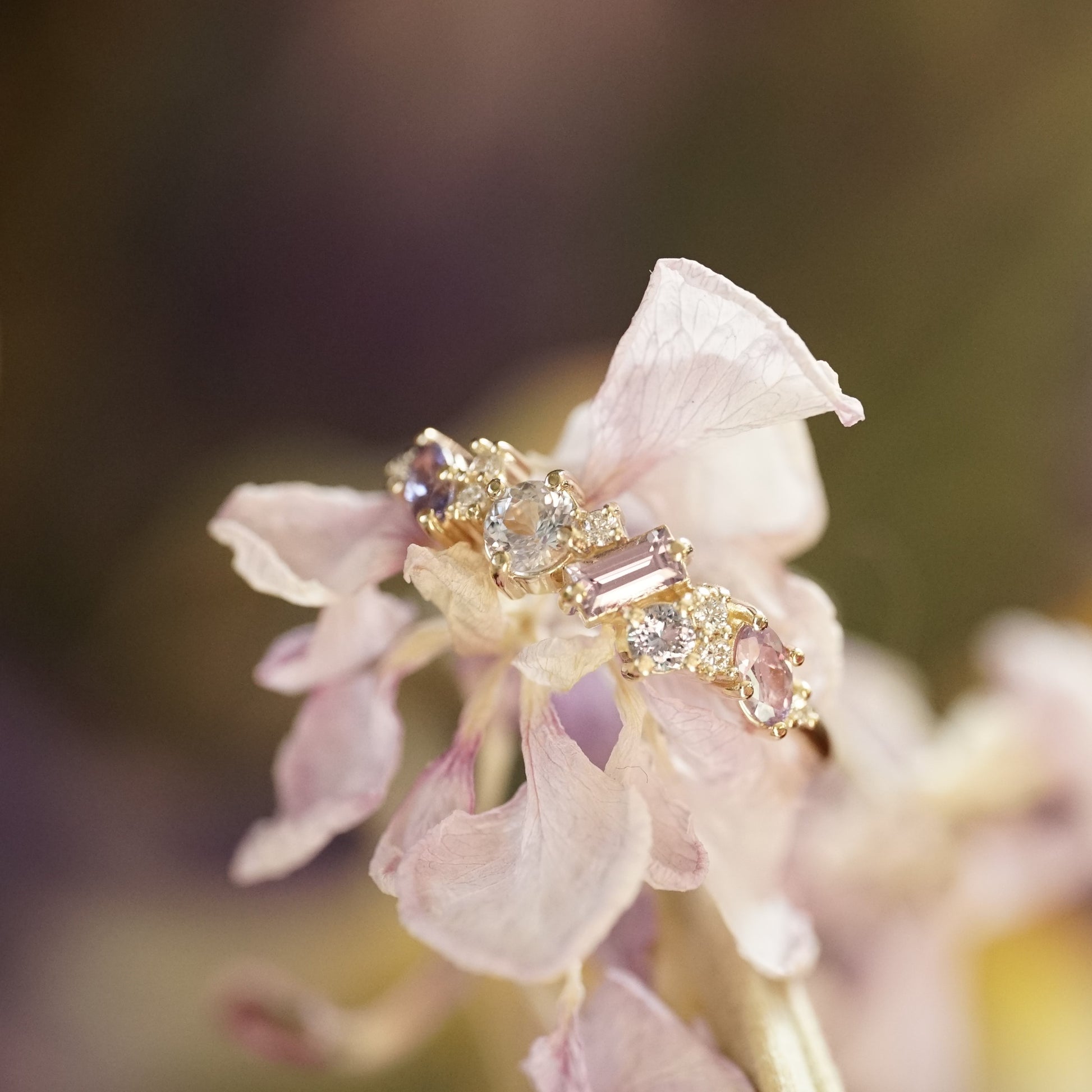 a yellow gold ring featuring purple and blue sapphires of mixed shapes and sizes are paired with round diamonds in a playful design. the ring rests on a very light pink flower